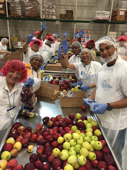 Volunteers at a food bank excitedly wave at the camera, they're packaging apples.